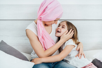 Cancer mother with a pink headscarf tenderly hugs her blonde daughter