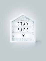 stay safe lettering on a lightbox with wooden house, concept of self quarantine as preventative measure against virus. inspiration quote, staying at home during pandemic and second wave of covid-19