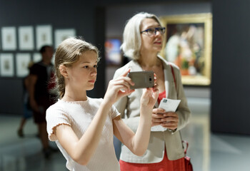 Fototapeta na wymiar Attentive tween girl and senior woman using phone for search info about painting in museum