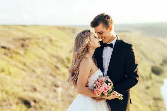 Portrait of young bride and groom enjoying romantic moments outside at sunset in beautiful summer day. Wedding couple. Standing face to face with the green hills on background.