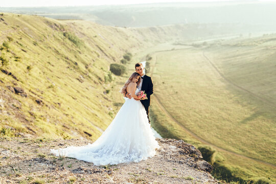 Full length body portrait of young bride and groom enjoying romantic moments outside at sunset in beautiful summer day. Wedding couple. Standing face to face with the green hills on background.