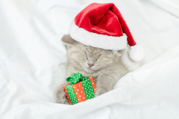 Funny kitten wearing red santa's hat sleeps under blanket with gift box. Top down view