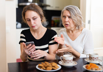 Upset mature mother with adult daughter using smartphone in kitchen interior