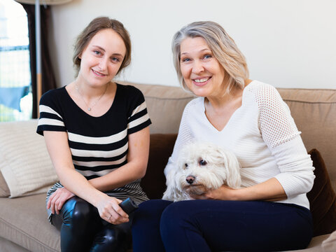 Positive family of elderly woman and adult girl with little dog snuggling on sofa, watching tv