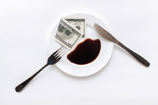 A Hundred-dollar Bill Lies In A White Plate Near A Fork, Knife And Fuel Oil, Top View. The Concept Of Petrodollars, Resource Extraction And Their Prices On The Exchange.