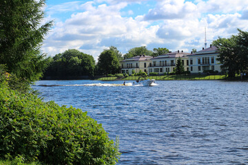 Obraz premium The girl is engaged in wakesurfing in the city canal against the background of low-rise buildings, trees and blue sky