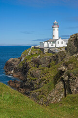 Fototapeta na wymiar The white painted lighthouse at Fanad Head, Donegal, Ireland stands on a cliff top above the blue Atlantic Ocean, a safety beacon for shipping in the dangerous coastal waters around the rocky shores.