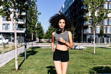 An athletic African-American young woman is jogging outdoors. A girl with an afro hairstyle in short sports bra and shorts runs on the street. Healthy lifestyle concept. Front view