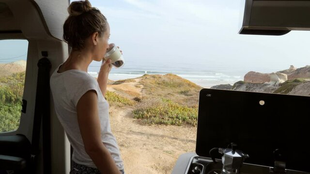 Young Woman Having A Fresh Cup Of Coffee In Her Camper Van Enjoys Coastal Views From Her Vehicle. Female Solo Traveller Living In Her Van Drinking Coffee In The Morning 