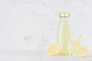 Fresh yellow lemon juice in glass bottle mock up with straw, wine glass, fruit slice on white wood table in light interior, template for packaging, advertising, design, branding product.