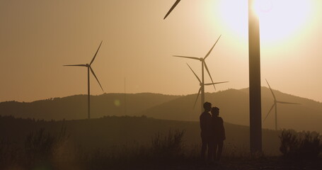 Two young men standing near the wind turbines pointing their hand forward. Silhouette shooting. Sunset