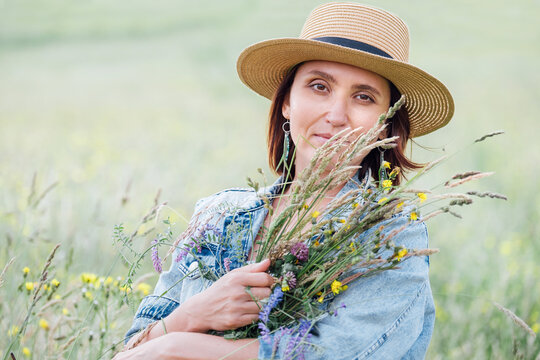 Portrait of a calm young woman with wildflowers bouquet. She dressed a jeans jacket, straw hat, and summer light dress. Natural people's beauty in the nature concept image.