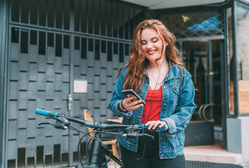 Portrait of Red curled long hair caucasian teen girl on the city street walking with bicycle using the smartphone with earphones. Natural people beauty urban life concept image.