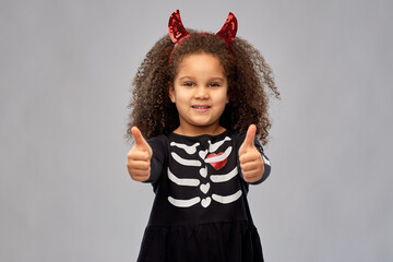 halloween, holiday and childhood concept - smiling african american girl in black costume dress and red devil's horns over grey background