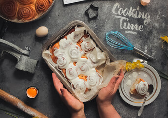 Homemade cinnamon rolls with white cream icing ,word cooking time made of white flour, blue whisk, egg shell on the grey dark concrete background, plate, notebook and pen, daffodil, hand try taste it