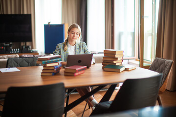 Young cute female student sitting with books and working at laptop. Back to school. Online education