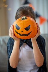 halloween, holiday and childhood concept - smiling girl in costume with jack-o-lantern at home