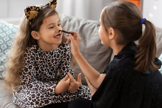 halloween, holiday and childhood concept - smiling little girls in party costumes doing face painting at home