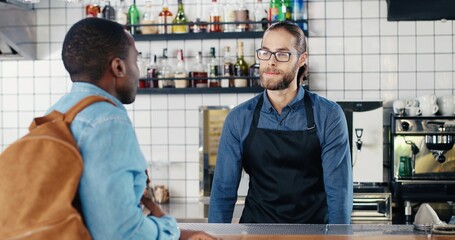 Caucasian male bartender taking order from African American guy tourist client at counter. Mixed-races. Man customer ordering drink and food at cafe from barrista. Multiethnic men. Asking for menu.