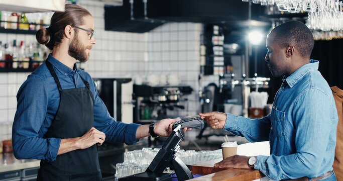 Joyful African American man client in cafe paying with credit card. Mixed-races. Caucasian bartender giving order and taking money. Multiethnic customer and barrista. Pay for coffee drink with paypass