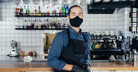 Portrait shot of young Caucasian handsome man waiter in mask and gloves standing at counter in cafe and looking at camera. Good-looking male barrista in apron at bar. Covid-19 quarantine concept.