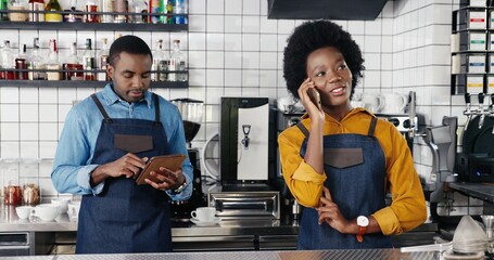 Pretty African American female waitress in apron standing in cafe at counter, talking on mobile phone. Man barrista tapping, texting and scrolling on tablet device on background. Speaking on cellphone