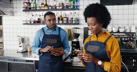 Beautiful African American female waitress in apron standing in cafe at counter and texting message on smartphone. Man barrista tapping and scrolling on tablet device. Sms on phone. Chatting online.