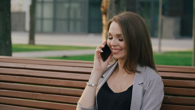 Happy brunette young woman talking on cellphone outdoors while sitting on bench in city park. Cheerful joyful lady finishes talking on the phone, hangs up and drinking coffee outside on sunny day.