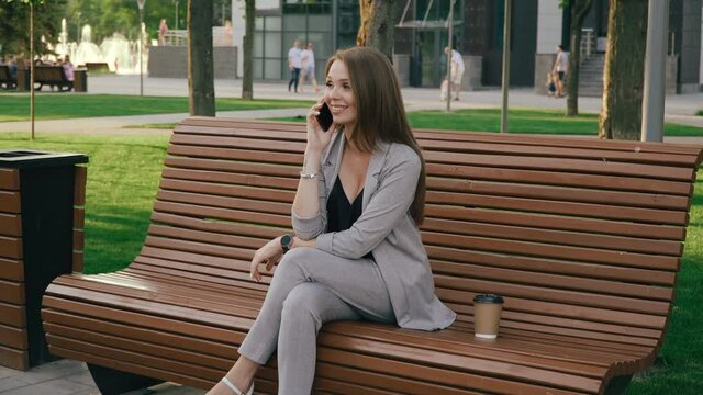 Happy brunette young woman talking on cellphone outdoors while sitting on bench in city park. Cheerful joyful lady finishes talking on the phone, hangs up and drinking coffee outside on sunny day.