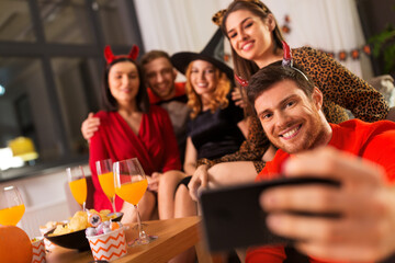 friendship, holiday and people concept - group of happy smiling friends in halloween costumes of vampire, devil, witch and cheetah taking selfie by smartphone at home party at night