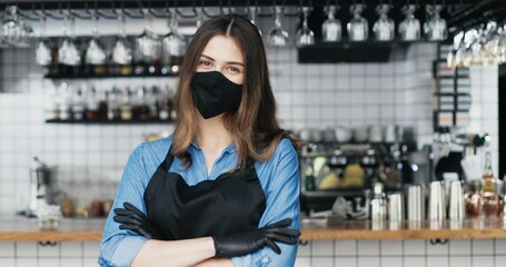 Portrait of Caucasian young beautiful woman barrista in mask and gloves standing at counter in bar, looking at camera and crossing hands. Waitress posing in cafe with drinks equipment on background.