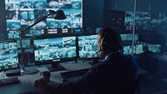 Male Officer Works on a Computer with Surveillance CCTV Video Footage in a Harbour Monitoring Center with Multiple Cameras on a Big Digital Screen. Employees Sit in Front of Displays with Big Data.