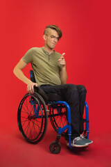 Pointing. Caucasian young disabled man's portrait isolated on red studio background. Beautiful male model in casual style. Concept of human emotions, facial expression, sales, ad, inclusion. Copyspace