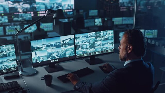 Male Officer Works on a Computer with Surveillance CCTV Video Footage in a Harbour Monitoring Center with Multiple Cameras on a Big Digital Screen. Employees Sit in Front of Displays with Big Data.