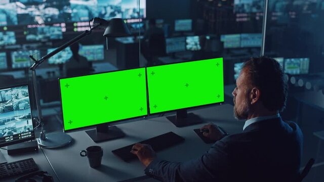 Male Officer Works on a Computer with Two Green Screen Mock Ups in Surveillance Center in a Harbour Monitoring Center with Multiple Cameras on a Big Digital Screen. Employees Sit in Front of Displays.
