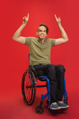 Pointing. Caucasian young disabled man's portrait isolated on red studio background. Beautiful male model in casual style. Concept of human emotions, facial expression, sales, ad, inclusion. Copyspace