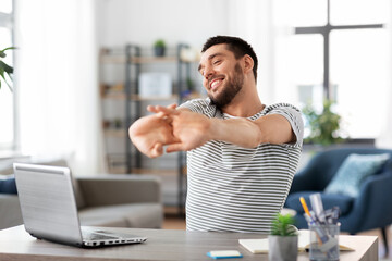 technology, remote job and business concept - happy smiling man with laptop computer stretching at...