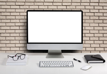 computer display for mockup in office interior on brick wall background. Work desk with keyboard, mouse, glasses, book.