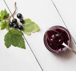 Currant homemade jam in glass jar on the white wooden table decorated with black currant and green leaves.