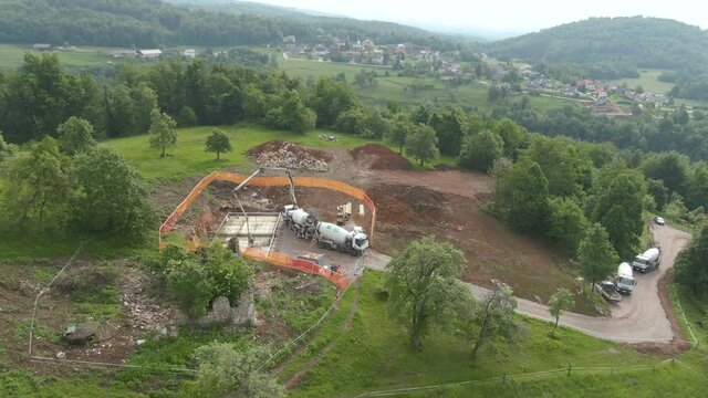 AERIAL: Big truck pours cement on the ground, creating the foundation of a house under construction. Flying above a busy construction site on a green hill in the countryside during cement pouring.