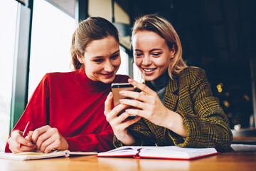 Positive female students watching live stream in blog on smartphone connected to 4G internet during exam preparation in cafe.Happy young women reading funny comments in social networks on telephone
