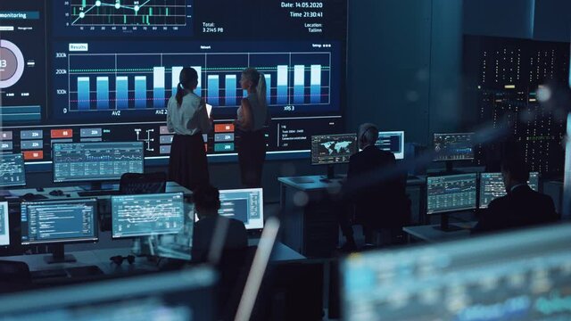 Two Software Engineers Talking Next to a Big Screen in a Modern Monitoring Office with Live Analysis Feed with Charts. Monitoring Room Big Data Scientists and Managers Sit in Front of Computers.