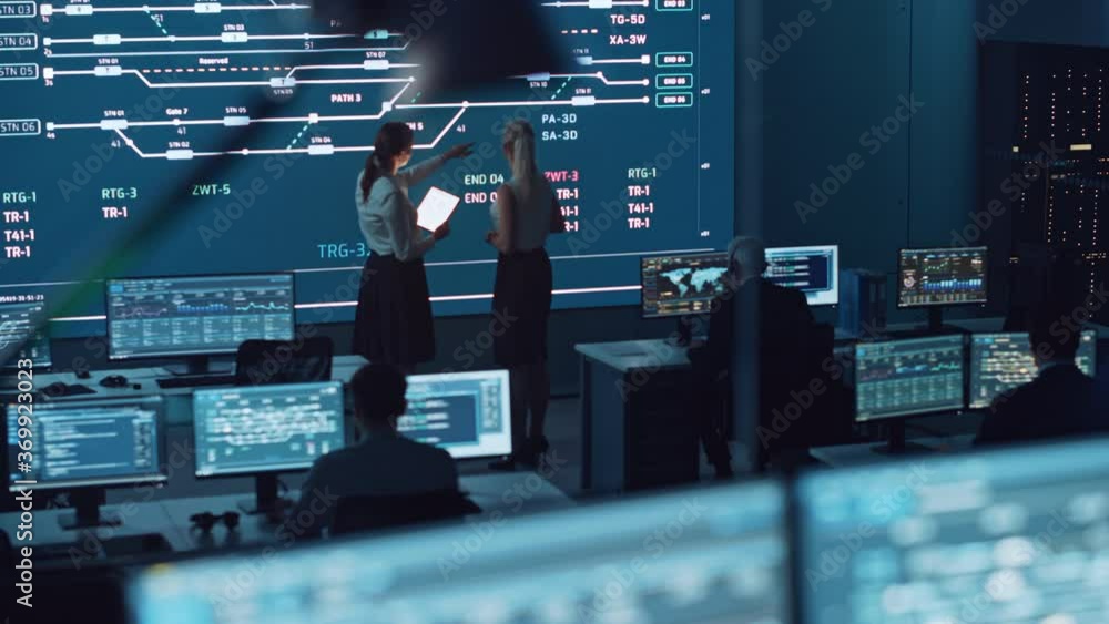 Poster Two Software Engineers Talking Next to a Big Screen in a Modern Monitoring Office with Live Analysis Feed with Charts. Monitoring Room Big Data Scientists and Managers Sit in Front of Computers.