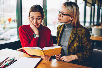 Shocked skilled student reading textbook with confused information during private lesson with qualified coach in coworking.Wondered hipster girl analyzing studying material together with tutor