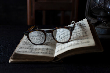 Old worn Glasses resting on an opened book with books and clock in background