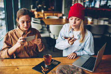 Addicted teenagers dressed in stylish casual wear ignoring live communication while making photos of tasty tea on modern smartphone for publication in social networks resting together in cafe interior