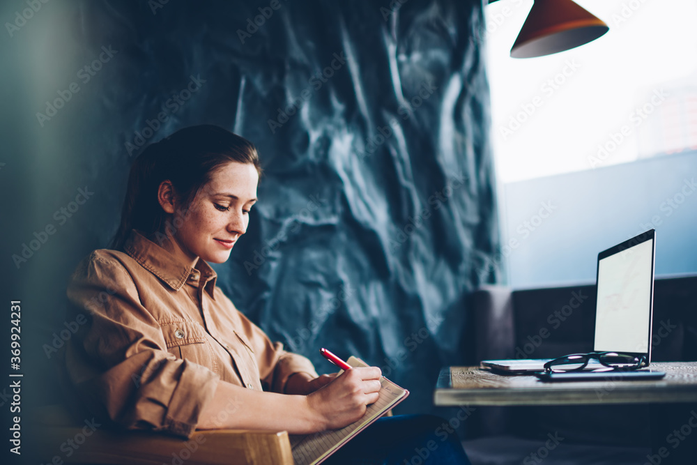 Poster creative student writing down text information in notebook sitting at modern laptop device with blan