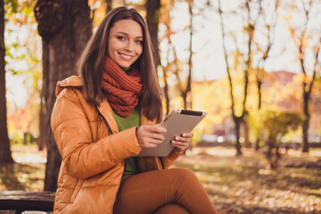 Photo of charming cute lady smiling sit bench park hold tablet waiting boyfriend browsing news feed share autumn pictures wear scarf green turtleneck orange windbreaker pants outdoors