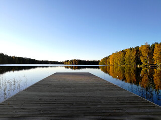 Colorful autumn landscape. A pier on the calm water of the lake. Yellow and red trees in the rays of the setting sun.