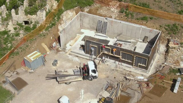 AERIAL: Flying around a construction crew laying sheets of metal wiring around the unfinished house walls. Group of contractors begin working on the basement floor of a modern house under construction